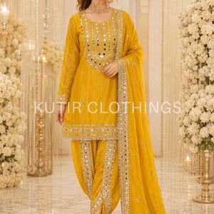 May include: A mustard yellow salwar kameez set featuring a long-sleeved tunic with intricate mirror work embroidery around the neckline and hem, paired with matching dhoti-style pants and a coordinating dupatta, all accented with silver mirror details.