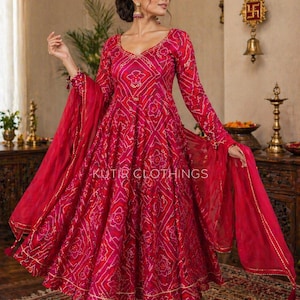 Puede incluir: Una mujer con un vibrante vestido rojo y rosa de estilo indio con un patrón geométrico. El vestido tiene mangas largas y una falda fluida, combinado con pantalones a juego y una bufanda transparente. El texto "KUTIR CLOTHINGS" es visible en el vestido.