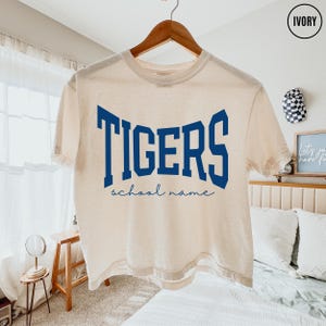 May include: An ivory short-sleeve t-shirt featuring the word TIGERS in large, bold, dark blue block lettering across the chest, with the words school name written in a smaller, cursive dark blue script font directly underneath the main text.
