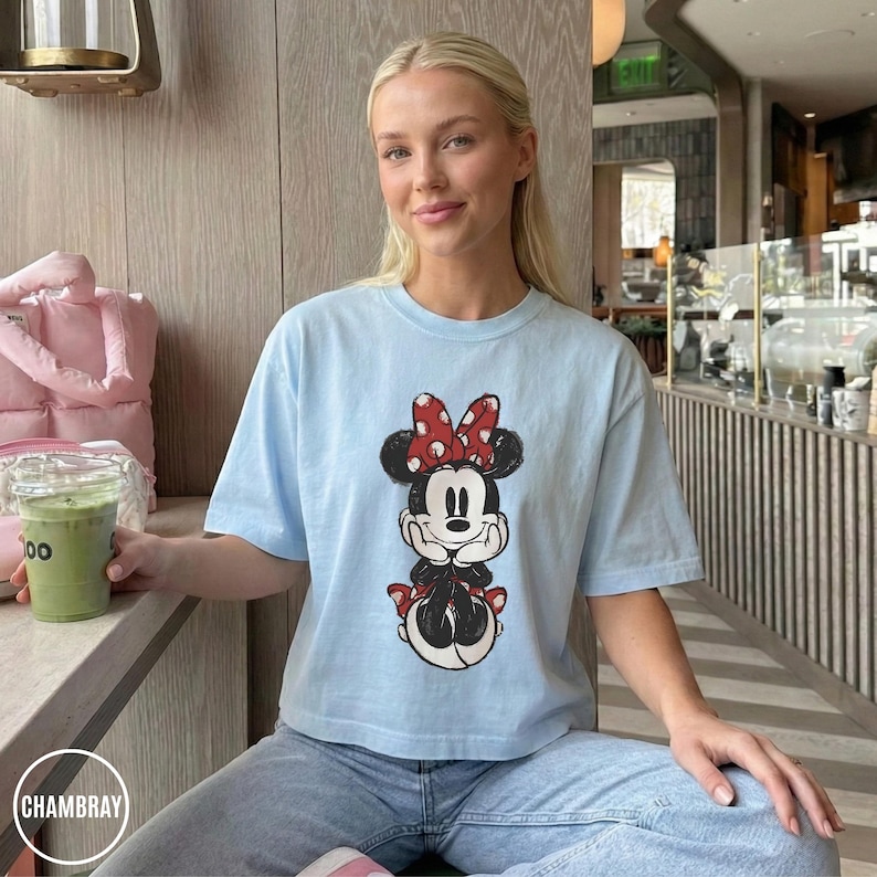 May include: Light blue t-shirt featuring a Minnie Mouse graphic. The cartoon character is depicted in black, red, and white. The shirt is paired with blue jeans. The word "CHAMBRAY" is visible in a white circle.