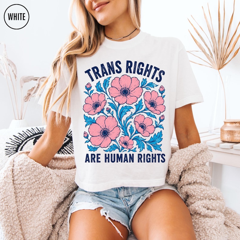 May include: A white t-shirt featuring a graphic print with the text "TRANS RIGHTS ARE HUMAN RIGHTS" in dark blue lettering. The design includes a central illustration of pink flowers with blue leaves and stems arranged in a square pattern.