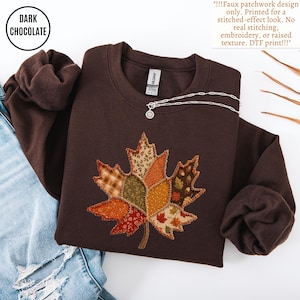 May include: A dark chocolate brown crewneck sweatshirt featuring a central faux patchwork maple leaf design. The leaf graphic consists of various autumn-toned patterns including plaid, floral, and acorn prints with a printed stitched-effect border.