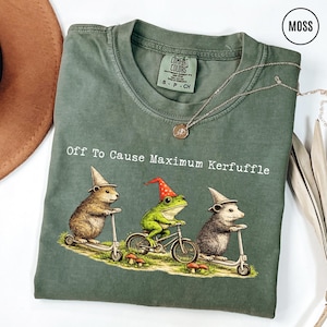 May include: Moss green t-shirt with the phrase "Off To Cause Maximum Kerfuffle." The graphic features a beaver on a scooter, a frog on a bicycle, and a possum on a scooter, all wearing hats. A silver necklace is draped across the shirt.