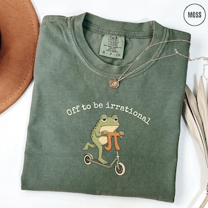 May include: Moss green t-shirt with the text "Off to be irrational" and a cartoon frog riding a scooter. The frog is green and tan, and the scooter is orange and gray. A gold necklace is draped across the shirt.