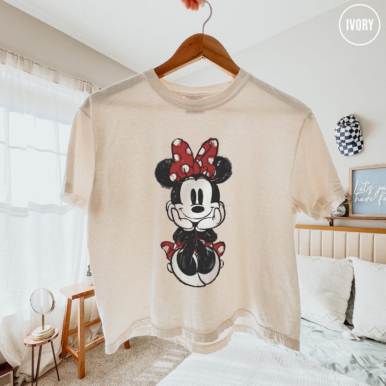May include: Cream-colored t-shirt featuring a graphic of Minnie Mouse. Minnie is depicted in black, red, and white, with a red polka-dot bow. The shirt is hanging on a wooden hanger.