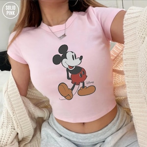 May include: A light pink crop top featuring a classic Mickey Mouse graphic. The cartoon character is depicted in his signature red shorts, brown shoes, and black ears. The top has short sleeves and the word "Disney" is printed on the bottom right.