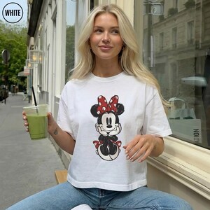 May include: White t-shirt featuring a Minnie Mouse graphic with a red polka dot bow. The cartoon character is depicted in a seated pose. The person is wearing blue jeans and holding a green beverage in a clear plastic cup.