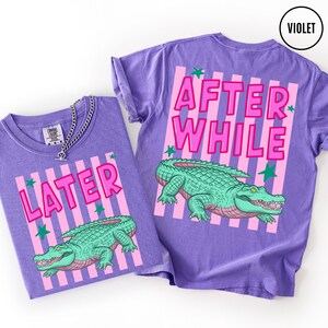 Puede incluir: Dos camisetas violetas con un dise&ntilde;o gr&aacute;fico. Una camiseta dice "LATER" y la otra "AFTER WHILE" en letras rosas. Ambas camisetas presentan una ilustraci&oacute;n de un caim&aacute;n verde con detalles rosas y rayas verticales rosas.