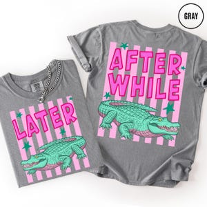 Puede incluir: Dos camisetas grises con dise&ntilde;os de rayas rosas y blancas. Una camiseta dice "LATER" y la otra "AFTER WHILE" en letras rosas. Ambas camisetas presentan una ilustraci&oacute;n de un caim&aacute;n verde.