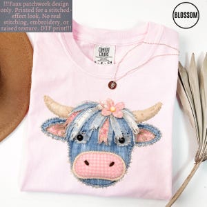 May include: A light pink t-shirt with a patchwork design of a Highland cow's face. The cow has blue denim-like hair, pink gingham snout, and a pink flower. A delicate necklace with a small pendant rests on the shirt.