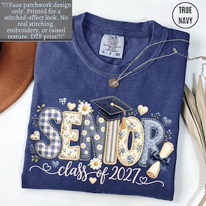 May include: A true navy blue t-shirt featuring a faux patchwork design with the text SENIOR class of 2027. The graphic includes a graduation cap, daisies, hearts, and a diploma tied with a blue ribbon. The print has a flat, stitched-effect look.