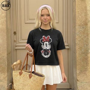 May include: Black t-shirt featuring a Minnie Mouse graphic with a red bow and polka dots. The t-shirt is paired with a white skirt and a straw tote bag with brown leather handles. A pink headband is worn.