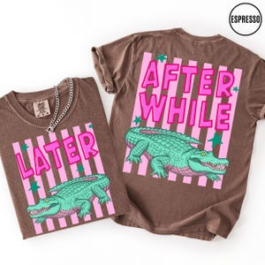 Puede incluir: Dos camisetas marrones con los textos "LATER" y "AFTER WHILE" en rosa, con un gr&aacute;fico de un cocodrilo verde. Las camisetas tienen un fondo a rayas rosas y detalles de estrellas verdes. Una camiseta tiene un collar de cadena plateada.