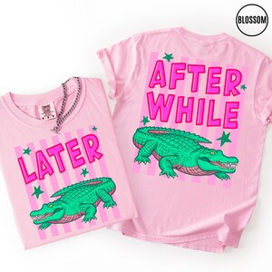Puede incluir: Dos camisetas rosas con las palabras "LATER" y "AFTER WHILE" en rosa brillante, junto con una ilustraci&oacute;n de un caim&aacute;n verde. Las camisetas tienen un fondo de rayas verticales y acentos de estrellas verdes. Una cadena de plata est&aacute; sobre una camiseta.