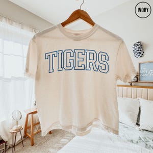 May include: An ivory short-sleeve t-shirt featuring the word TIGERS printed in a bold, blue, collegiate-style font across the chest. The garment has a crew neckline and a standard fit, displayed on a wooden hanger against a neutral background.