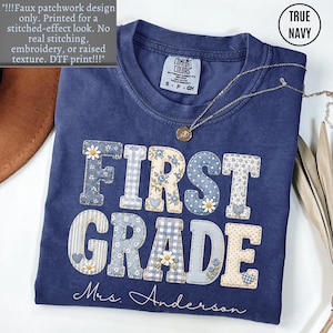 May include: A true navy blue t-shirt featuring a faux patchwork design with the text "FIRST GRADE" and "Mrs. Anderson" in white script. The letters have various blue and cream floral, gingham, and polka dot patterns with small daisy accents.