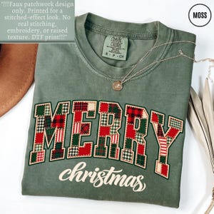 May include: A moss green t-shirt featuring the word MERRY in a faux patchwork design with red, green, and white plaid patterns, and the word christmas in white script below. The shirt has a Comfort Colors label and a small gold pendant necklace.