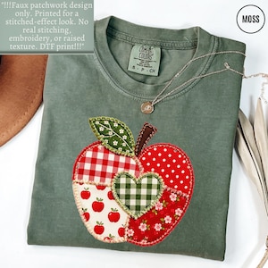 May include: A moss green t-shirt featuring a large, central apple graphic with a faux patchwork design. The apple is composed of various red and white patterns, including gingham, polka dots, and floral prints, with a green gingham heart.
