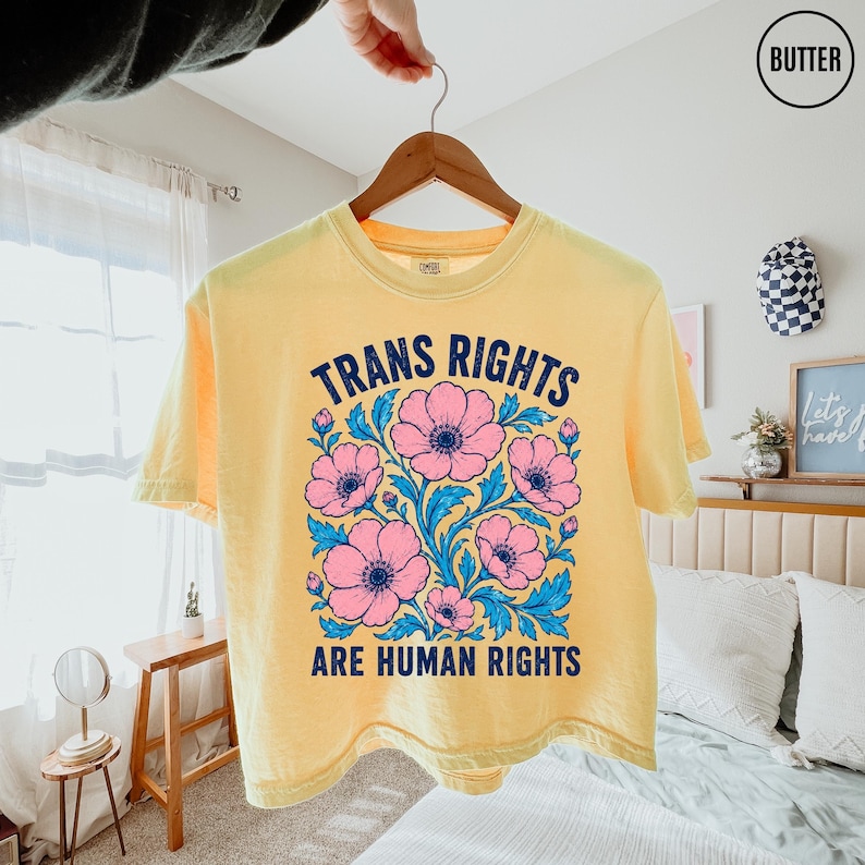May include: A pale yellow short-sleeve t-shirt featuring a graphic print of pink flowers with blue leaves. The text "TRANS RIGHTS ARE HUMAN RIGHTS" is printed in dark blue capital letters above and below the floral illustration.