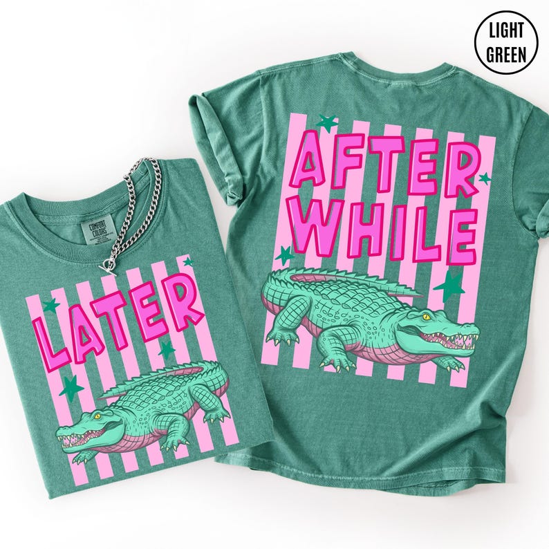 Puede incluir: Dos camisetas verde claro con dise&ntilde;os de rayas rosas y las palabras "LATER" y "AFTER WHILE" en letras rosas. Cada camiseta presenta una ilustraci&oacute;n de un caim&aacute;n verde con acentos rosas. Las camisetas son ropa casual.