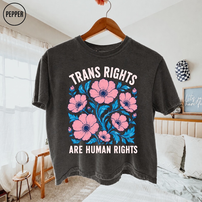 May include: A charcoal gray short-sleeve t-shirt featuring a graphic print of pink flowers with blue leaves. The text "TRANS RIGHTS ARE HUMAN RIGHTS" is printed in white capital letters above and below the floral design on the front of the shirt.