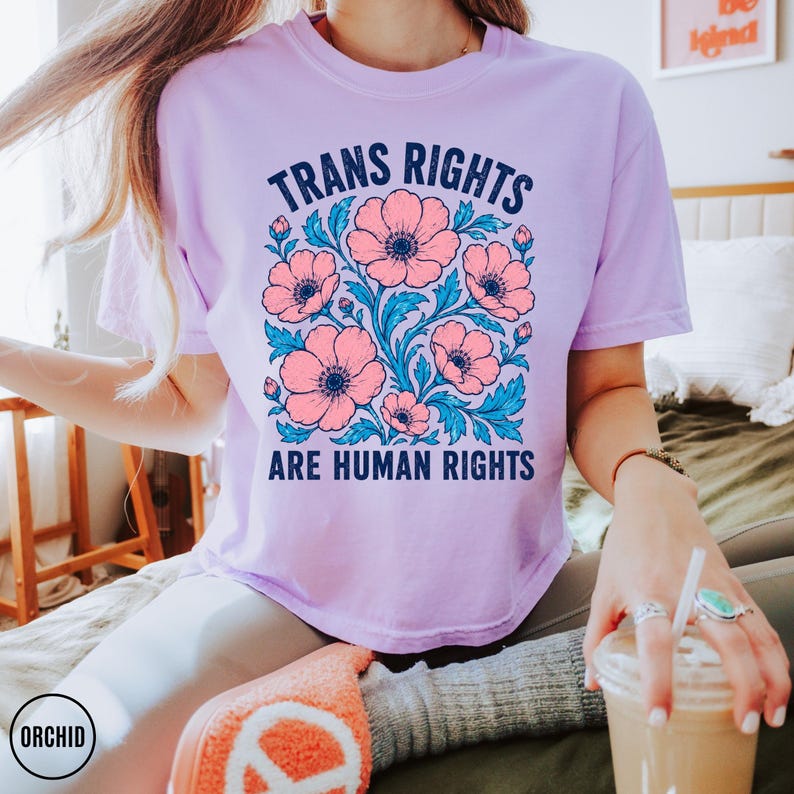 May include: A light purple short-sleeve t-shirt featuring a central graphic of pink flowers with blue leaves. The text "TRANS RIGHTS ARE HUMAN RIGHTS" is printed in dark blue capital letters above and below the floral illustration.