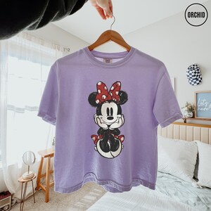 May include: A lavender t-shirt featuring a Minnie Mouse graphic. The cartoon character is depicted in black and white with a red polka-dot bow and matching accents. The shirt is hanging on a wooden hanger.