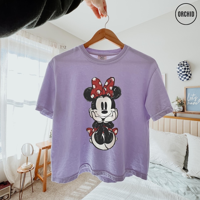 May include: A lavender t-shirt featuring a Minnie Mouse graphic. The cartoon character is depicted in black and white with a red polka-dot bow and matching accents. The shirt is hanging on a wooden hanger.