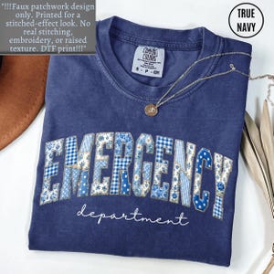 May include: A navy blue t-shirt with the word "EMERGENCY" in a patchwork design, and the word "department" below it. The shirt has a round neckline and short sleeves. A gold necklace with a pendant is on the shirt.