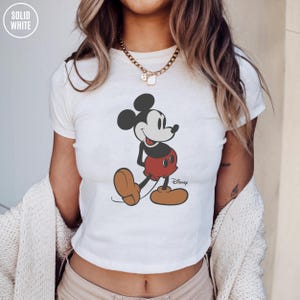 May include: White t-shirt featuring a classic Mickey Mouse graphic. The cartoon character is depicted in black, red, and brown. The Disney logo is printed below the character. The shirt has a crew neck and short sleeves.
