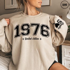 May include: A sand-colored crewneck sweatshirt featuring the black text 1976 Limited Edition on the front. The left sleeve displays a black heart icon with the text Chapter 5. The design has a distressed, vintage-inspired texture throughout.
