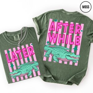 Puede incluir: Dos camisetas verde musgo con un dise&ntilde;o de rayas rosas y blancas. Una camiseta dice "LATER" y la otra "AFTER WHILE" en letras rosas. Cada camiseta presenta un gr&aacute;fico de un caim&aacute;n turquesa.