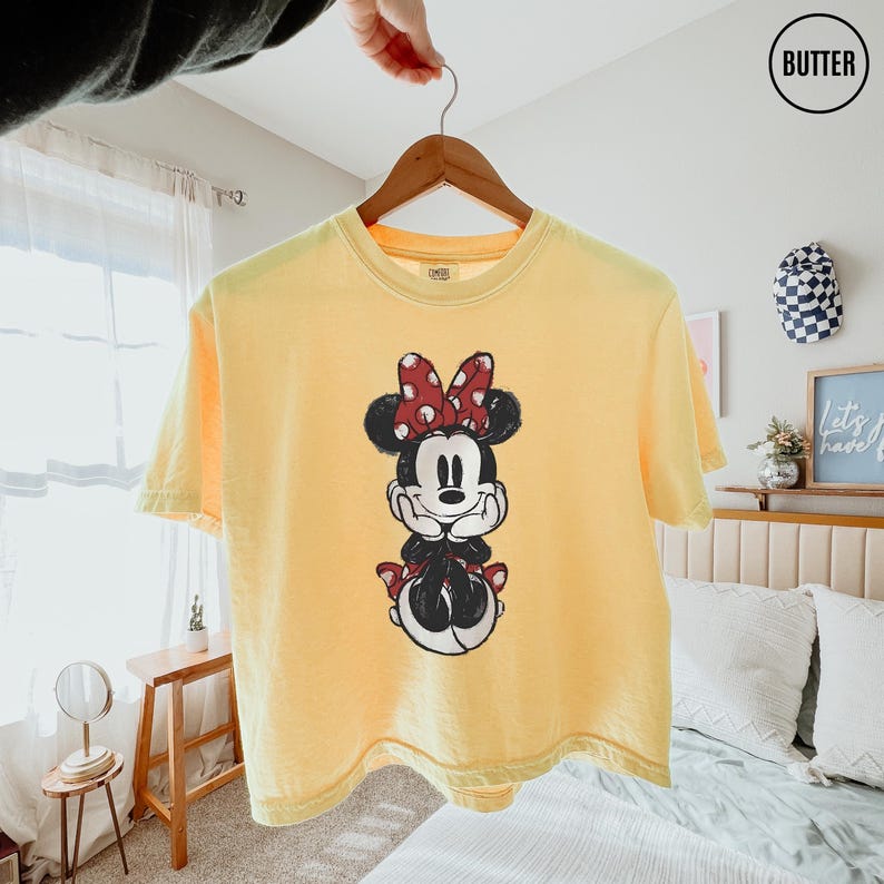 May include: Yellow t-shirt featuring a Minnie Mouse graphic. The graphic shows Minnie Mouse with a red polka dot bow, black ears, and a red polka dot bow on her shoes. The shirt is hanging on a wooden hanger.
