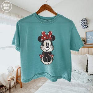 May include: A turquoise t-shirt featuring a graphic of Minnie Mouse with a red polka dot bow. The cartoon character is in a seated pose with her hands under her chin. The shirt is hanging on a wooden hanger.