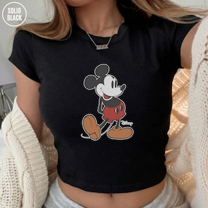 May include: Black ribbed crop top featuring a classic Mickey Mouse cartoon graphic. The Disney character is depicted in his iconic red shorts, brown shoes, and white gloves. The top has a crew neck and short sleeves. The words "SOLID BLACK" are in the top left corner.