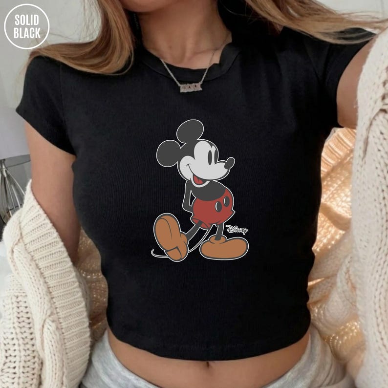May include: Black ribbed crop top featuring a classic Mickey Mouse cartoon graphic. The Disney character is depicted in his iconic red shorts, brown shoes, and white gloves. The top has a crew neck and short sleeves. The words "SOLID BLACK" are in the top left corner.