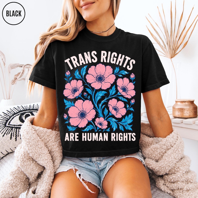 May include: A black short-sleeve t-shirt featuring a graphic print of pink flowers with blue leaves. The text "TRANS RIGHTS ARE HUMAN RIGHTS" is printed in white capital letters above and below the floral design in a distressed font style.