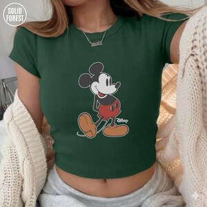 May include: Dark green crop top featuring a classic Mickey Mouse graphic. The cartoon character is depicted in black, white, red, and brown. The top has short sleeves and a ribbed texture. The Solid Forest logo is visible in the upper left corner.