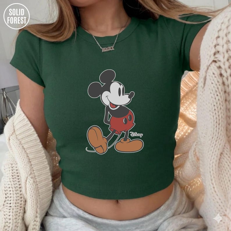 May include: Dark green crop top featuring a classic Mickey Mouse graphic. The cartoon character is depicted in black, white, red, and brown. The top has short sleeves and a ribbed texture. The Solid Forest logo is visible in the upper left corner.