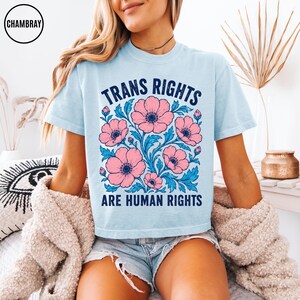 May include: A light blue t-shirt featuring a graphic print of pink flowers with blue leaves. The text "TRANS RIGHTS ARE HUMAN RIGHTS" is printed in dark blue capital letters above and below the floral design on the front of the garment.