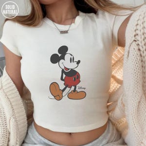 May include: Cream-colored crop top featuring a graphic of Mickey Mouse in his classic pose. The cartoon character is depicted in black, red, and tan colors. The top has the text "SOLID NATURAL" in a circle at the top left and the Disney logo at the bottom right.
