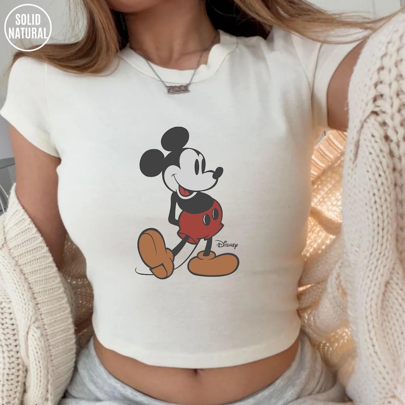 May include: Cream-colored crop top featuring a graphic of Mickey Mouse in his classic pose. The cartoon character is depicted in black, red, and tan colors. The top has the text "SOLID NATURAL" in a circle at the top left and the Disney logo at the bottom right.