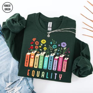 May include: Forest green sweatshirt with a colorful graphic of books with flowers and the words "LOVE, PEACE, INCLUSION, KINDNESS, CHANGE, UNITY, DIVERSITY, COMPASSION, JUSTICE" and the word "EQUALITY". A silver necklace is also visible.