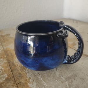 Könnte beinhalten: Ein runder Keramikbecher mit einer tiefblauen, glänzenden Glasur in gesprenkelter Optik. Der Henkel ist wie ein Tentakel geformt, mit sichtbaren Saugnäpfen an der Außenkante, und erstreckt sich seitlich vom abgerundeten Gefäßkörper.