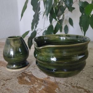 Könnte beinhalten: Zweiteiliges Matcha-Teeset aus Keramik mit glänzender, dunkelgrüner Glasur. Das Set besteht aus einem kleinen, abgerundeten Schneebesenhalter und einer größeren Schale mit Ausguss. Die Teile sind handgefertigt und haben eine rustikale, organische Ästhetik.