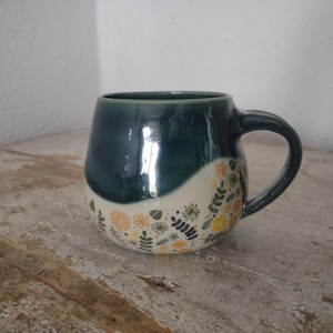 Könnte beinhalten: Ein Keramikbecher mit abgerundeter Form, der eine glänzend dunkel-petrolfarbene Glasur im oberen Bereich und eine cremefarbene Basis mit einem wiederkehrenden Muster aus kleinen gelben und grünen Blumen, Blättern und Zweigen aufweist.
