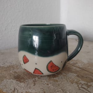 Könnte beinhalten: Ein Keramikbecher mit einem glänzend dunkelgrünen oberen Teil und einem cremefarbenen unteren Teil. Die cremefarbene Basis zeigt ein handgemaltes Muster aus roten Wassermelonenstücken mit schwarzen Kernen. Der Becher hat einen runden Körper.