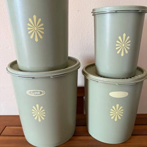 May include: A set of four sage green plastic canisters with matching lids. Each canister features a yellow starburst floral design. One canister has the word FLOUR printed in a yellow oval label on the front, while another has a blank yellow oval.