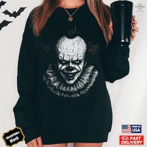 May include: Black Champion sweatshirt featuring a detailed, grayscale image of a scary clown's face. The clown has a white face, red lips, and a ruffled collar. The sweatshirt has the text "Champion Black" on the bottom left.