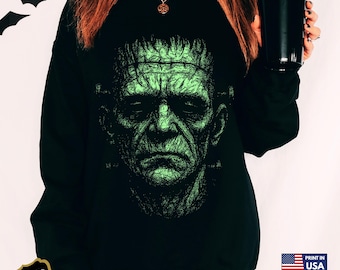 May include: Black Champion sweatshirt featuring a green and black graphic of Frankenstein's monster. The design is a detailed illustration of the monster's face. The sweatshirt has a crew neck and long sleeves. The text "Print in USA" is visible.